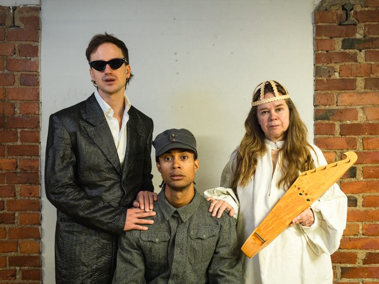 Miesoletettu puvussa ja aurinkolaseissa vasemmalla, keskellä istuu miesoletettu sotilaspuvussa, vasemmalla seisoo henkilö Väinämöisen asussa ja kantele käsissään.