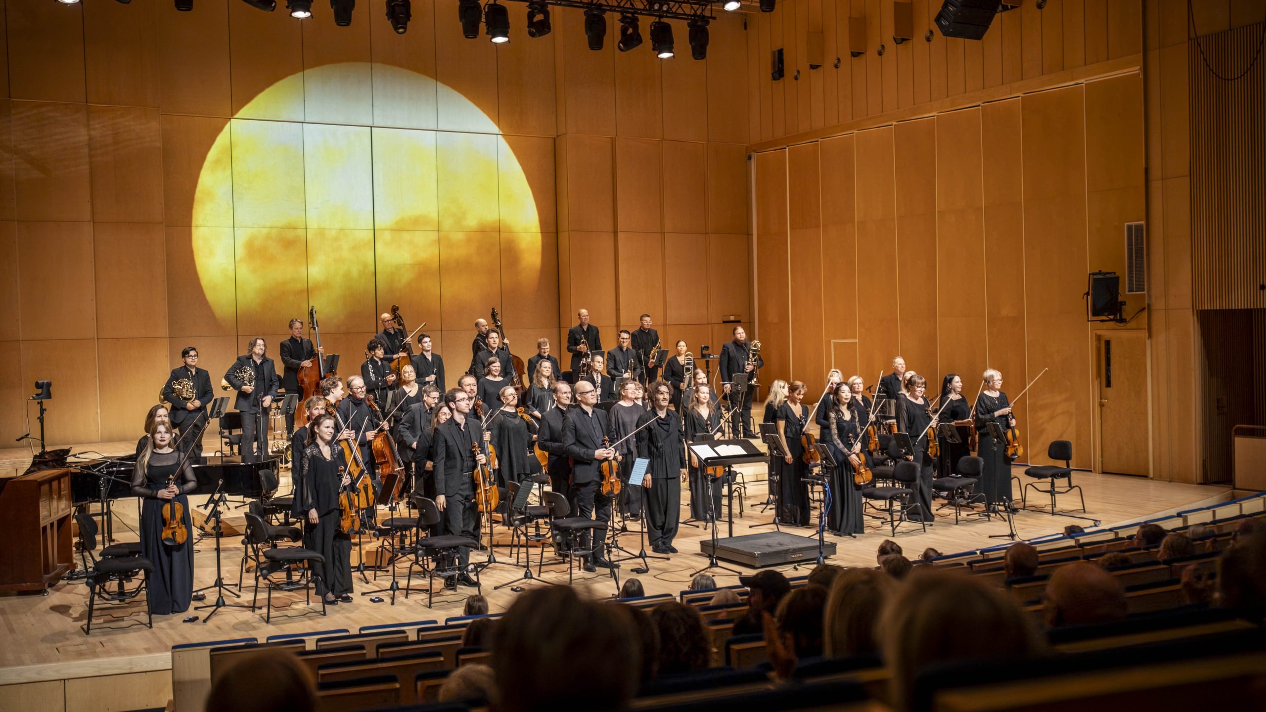 Tapiola Sinfoniettan orkesteri lavalla ottamassa vastaan suosionosoituksia. Taustalla heijastettuna kuu pilvimaisemassa.