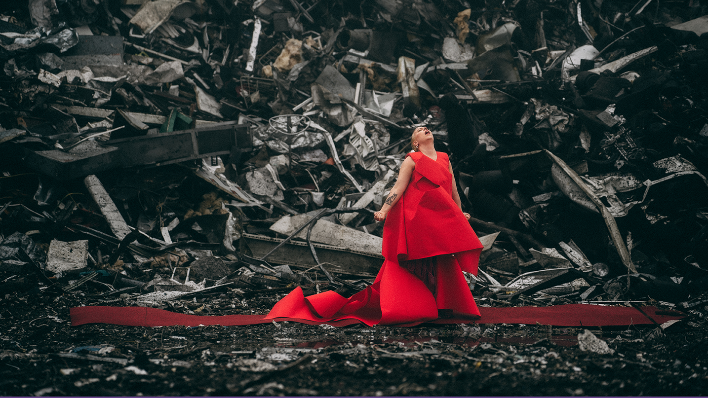 Muusikko Aili Järvelä punaisessa gaalamekossa tummanpuhuvan romukasan edessä.