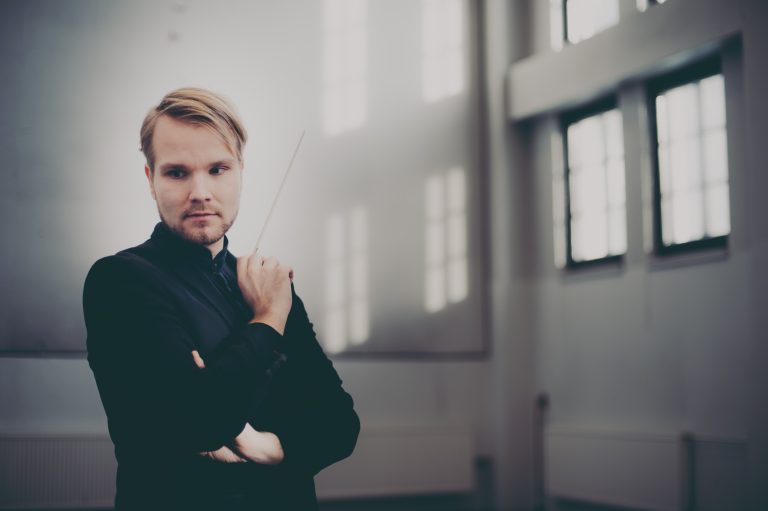 Henkilö seisoo suurten ikkunoiden valossa kapellimestarin puikko kädessään.