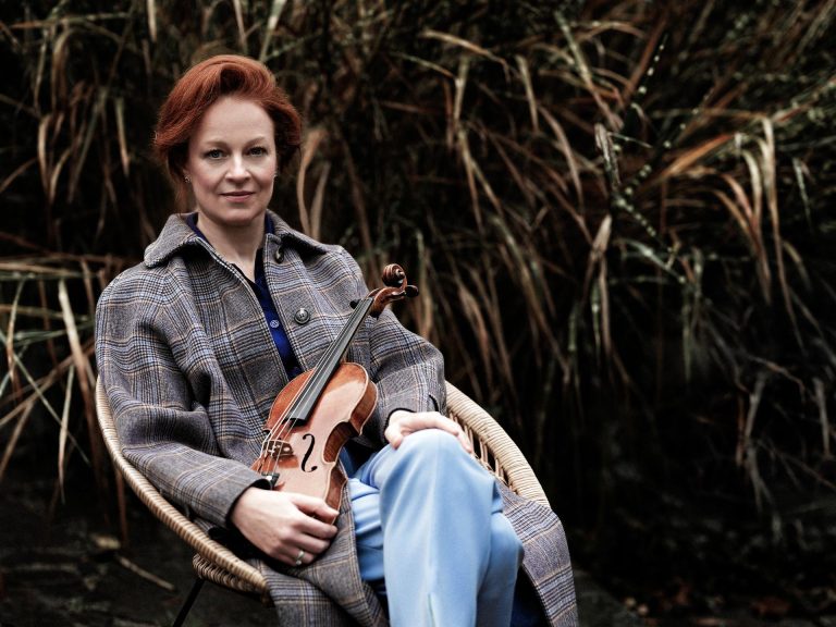 Pitkätakkinen nainen istuu tuolilla viulu sylissään.