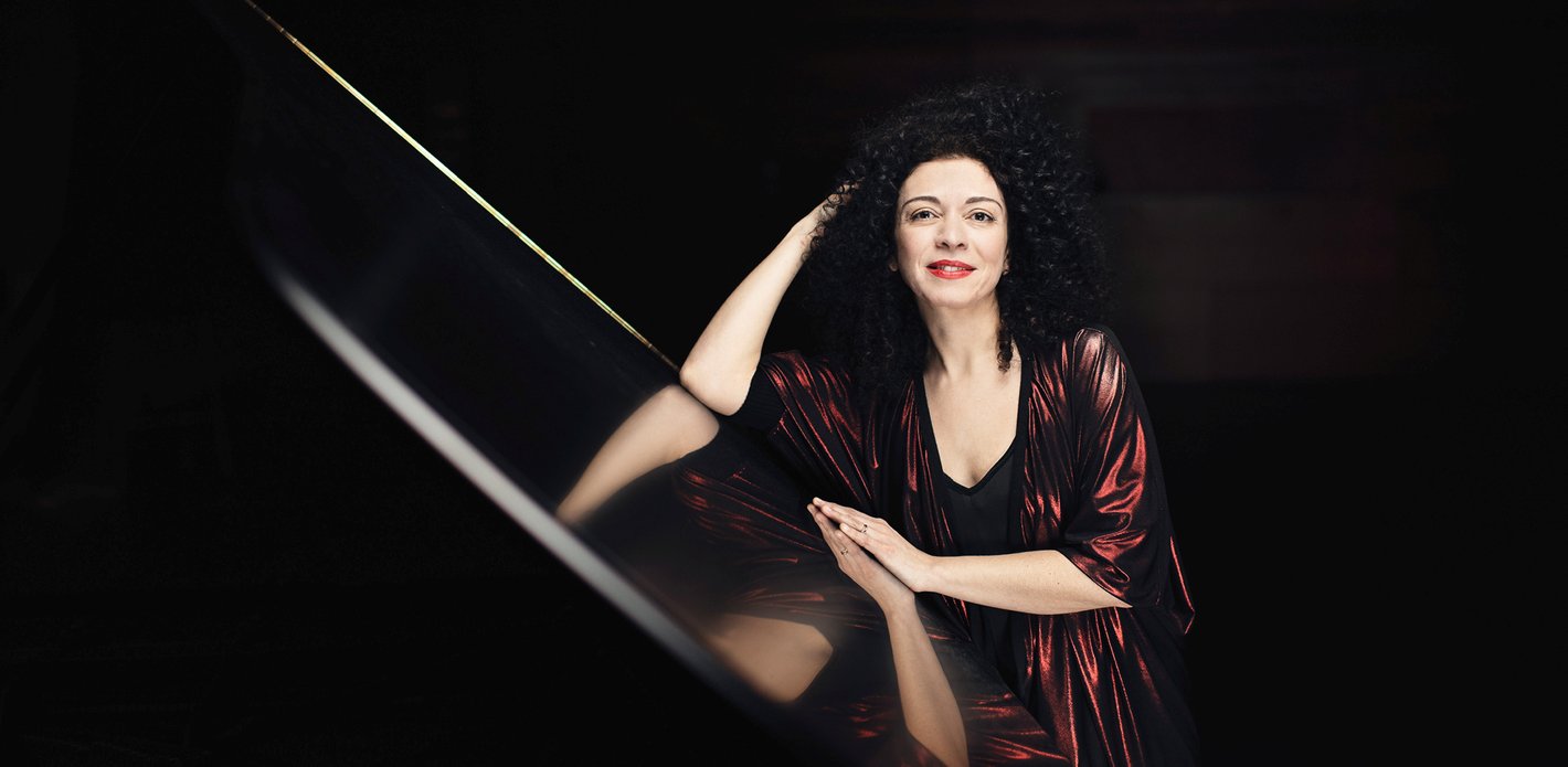 Tummahiuksinen nainen istuu pianon ääressä nojaten pianoon.