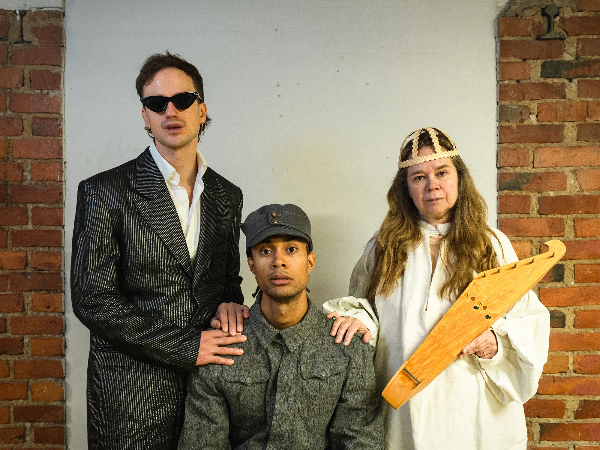 Miesoletettu puvussa ja aurinkolaseissa vasemmalla, keskellä istuu miesoletettu sotilaspuvussa, vasemmalla seisoo henkilö Väinämöisen asussa ja kantele käsissään.