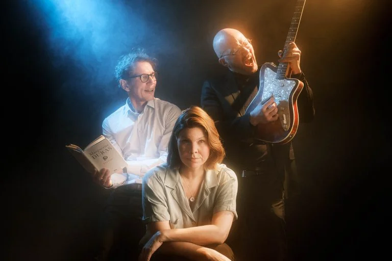 Kirjailija Juha Itkonen istuu kirja kädessään ja katsoo kitaristi Marzi Nymanin soittoa iloisena, keskessä istuu näyttelijä Eeva Soivio katsoen suoraan kameraan.