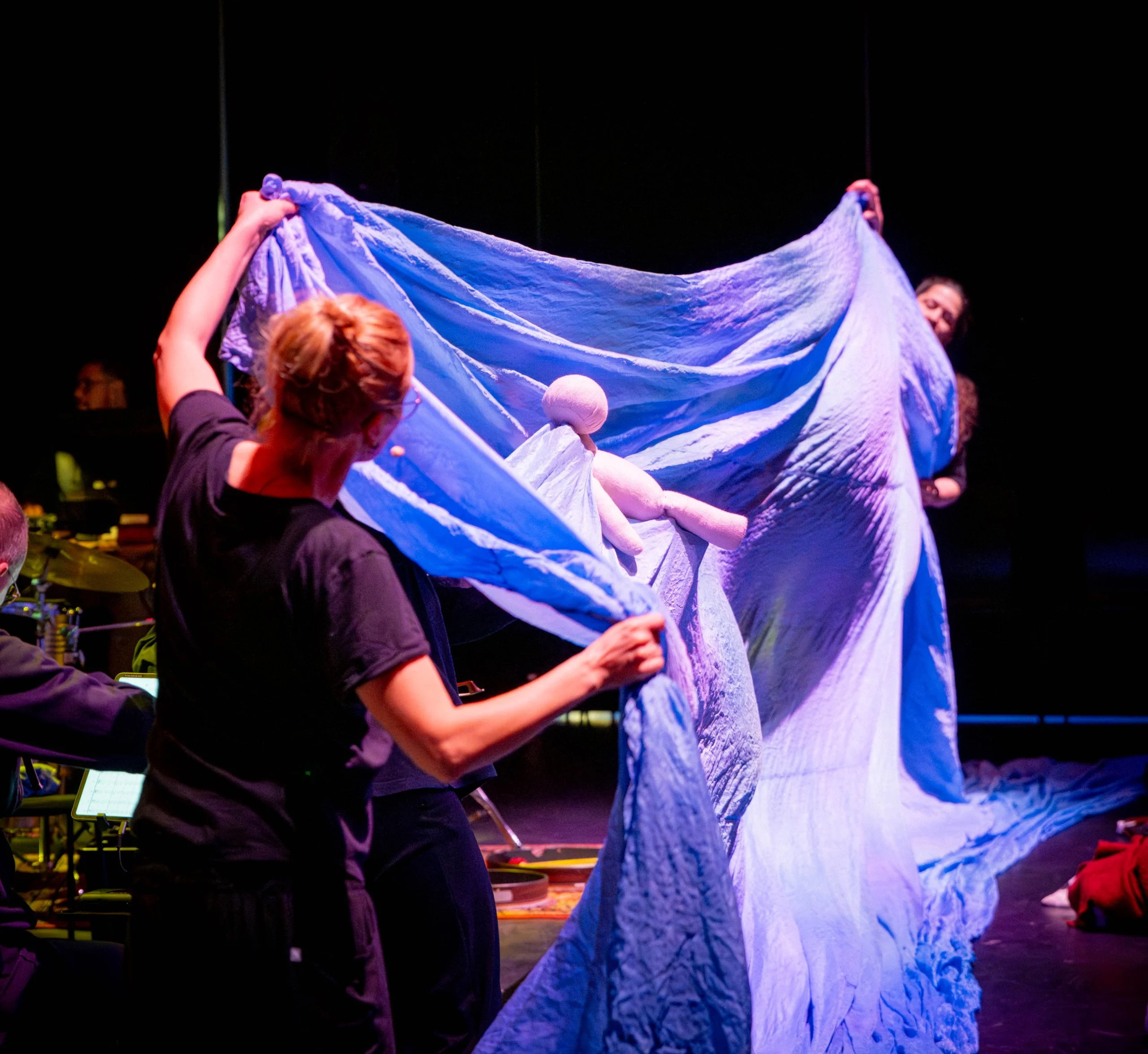 Kaksi henkilöä pitelee vaaleansinistä lakanaa, jonka päällä makaa pieni, yksinkertainen nukke.